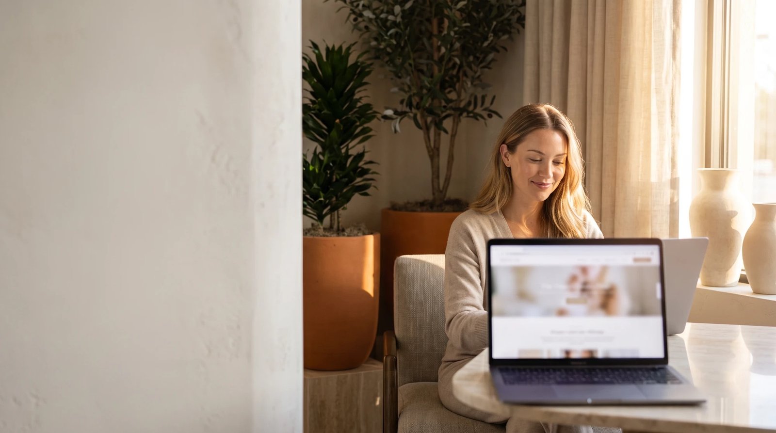 A small business owner smiling at her laptop in a bright room with terracotta planters, linen curtains, and cream vases